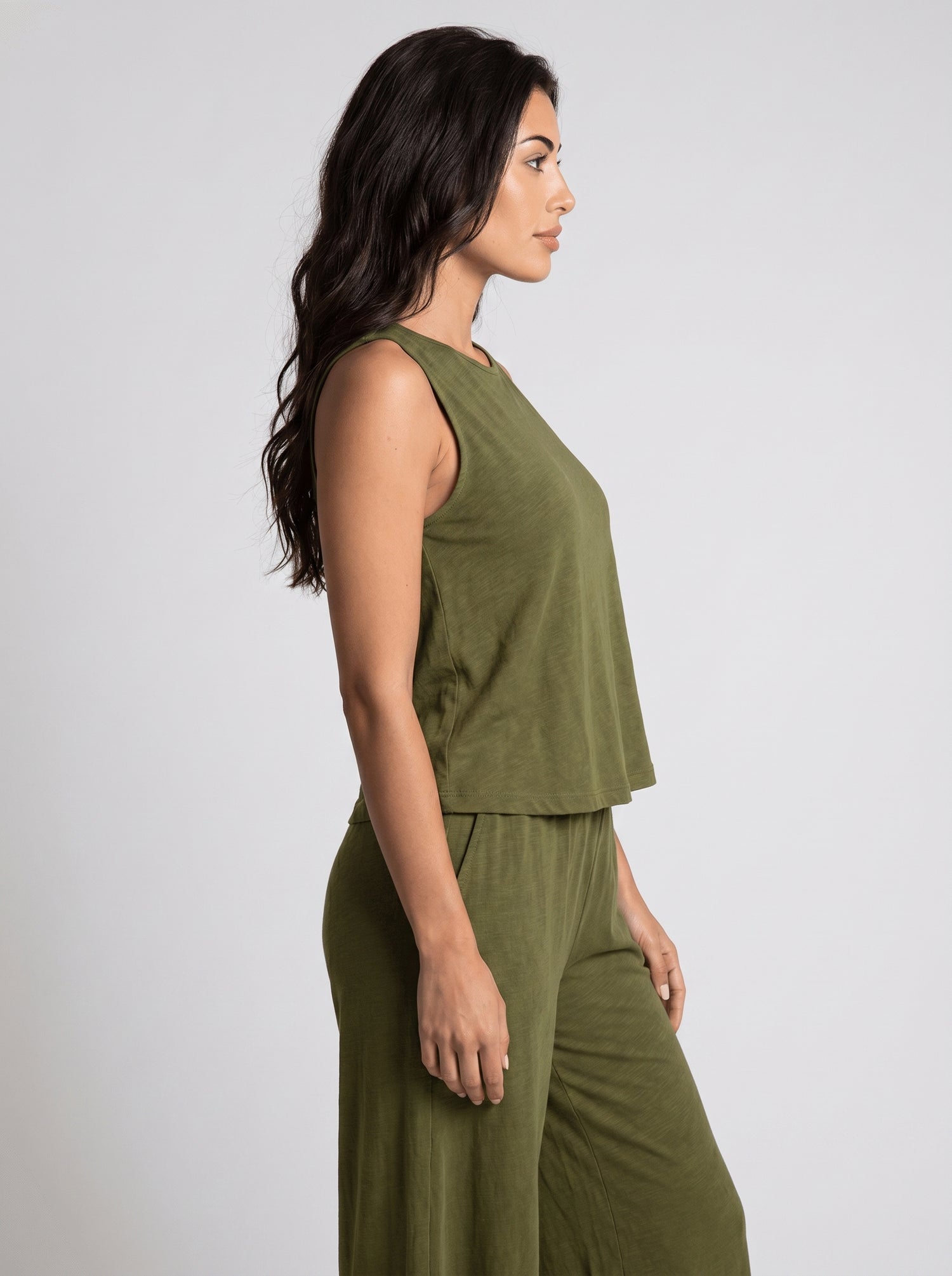 Side profile of woman wearing an olive green sleeveless crop top and matching wide-leg pants, arms relaxed, neutral studio background