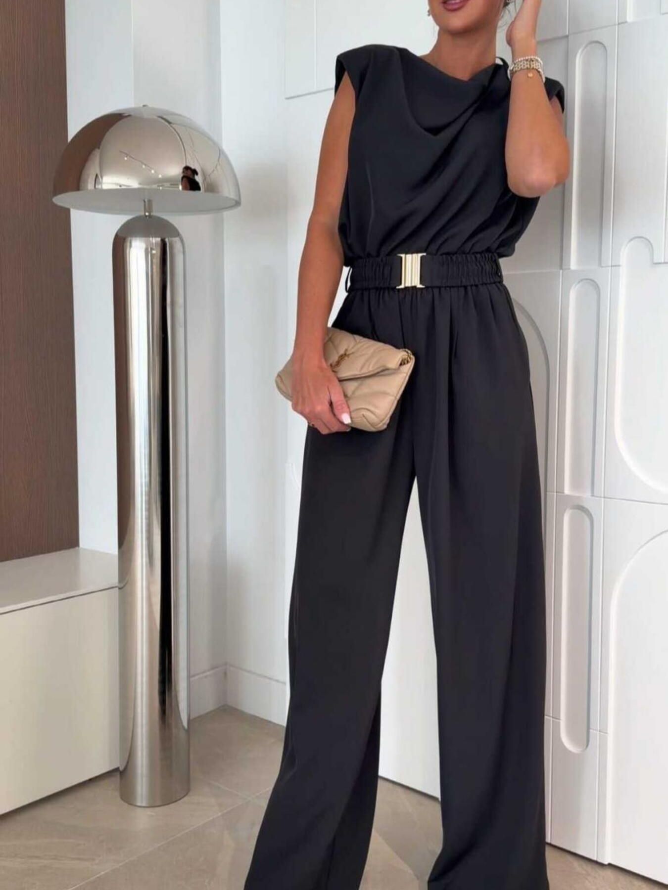A woman wearing a black sleeveless wide-leg jumpsuit with padded shoulders, a draped cowl neckline, ruched elastic waistband with a gold metal clasp, and flowing wide-leg trousers. She is styled with a beige quilted YSL clutch and a pearl bracelet, with one hand raised to her hair. Three-quarter front view.